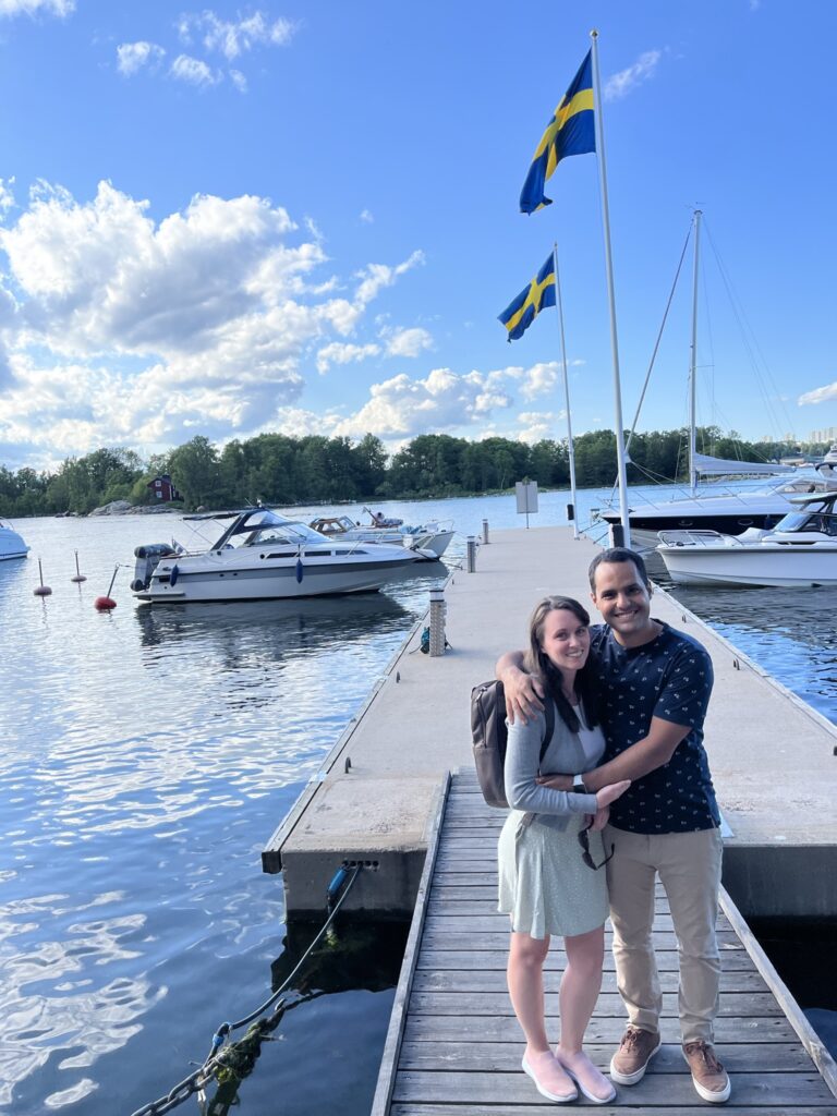 Couple smiling and embracing on a wooden dock by the water, with Swedish flags waving in the background, surrounded by boats and a bright blue sky with fluffy clouds.