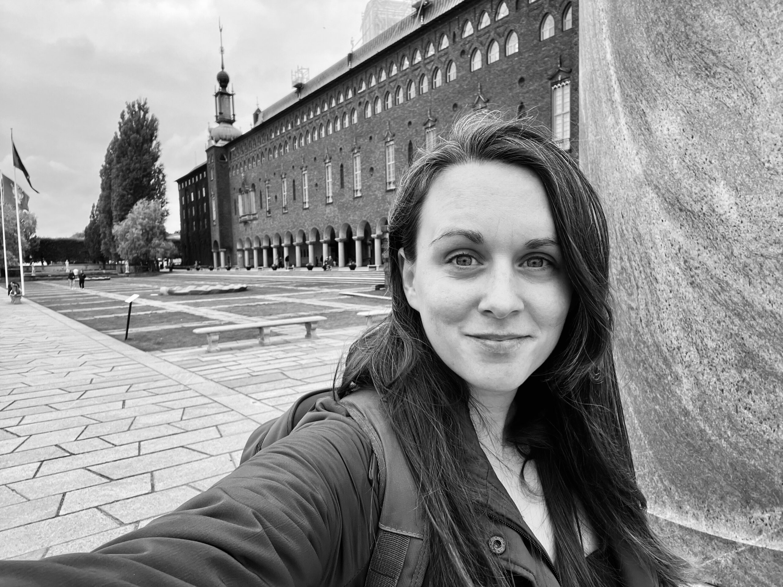 A black-and-white photo of a woman standing in front of the iconic Stockholm City Hall in Sweden. She smiles warmly at the camera, with the building's distinct red brick facade, arches, and spire visible in the background. The open courtyard and a hint of Swedish flags add to the scene's cultural significance.