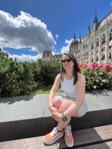 Woman smiling in front of the Hungarian Parliament Building, surrounded by colorful flowers under a bright blue sky.