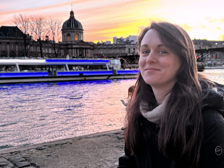 Person smiling next to the Seine River with a vibrant Paris sunset sky in the background.