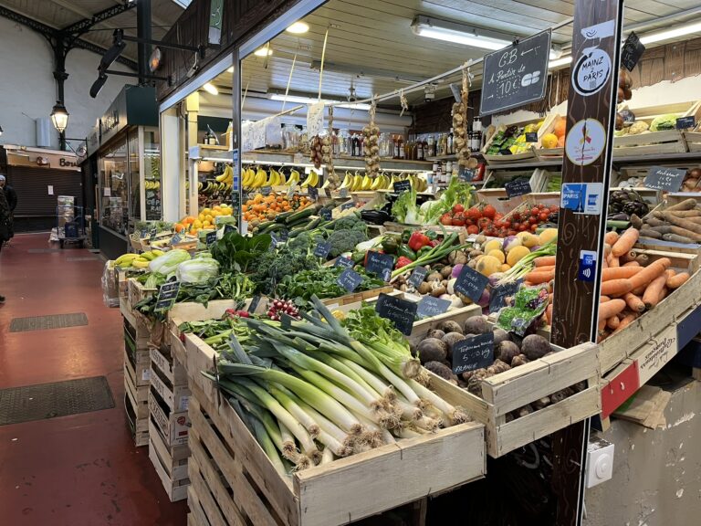 Colorful vegetable stand inside a French weekend market building, featuring fresh produce like tomatoes, carrots, and leafy greens.