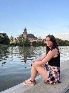 Woman sitting by the riverbank with Vajdahunyad Castle in the background, separated by the water and framed by peaceful scenery in Budapest’s City Park.