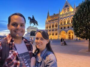 Couple smiling with the Hungarian Parliament Building illuminated in the background just after sunset.