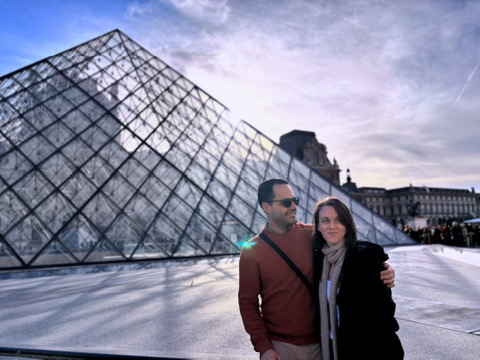 Couple standing in front of the Louvre Museum with the iconic glass pyramid in the background on a sunny day in Paris.
