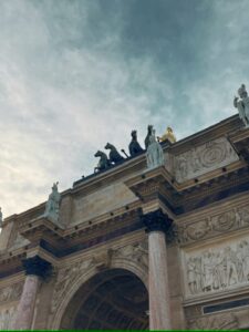 Partial view of the Arc de Triomphe du Carrousel near the Louvre, with horse statues on top and a bright sky in the background.