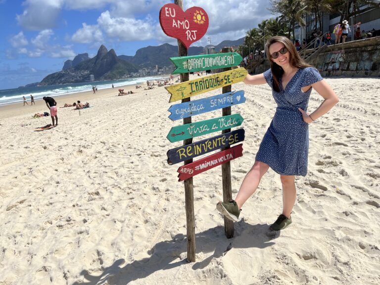 Woman in a blue dress striking a playful pose in front of a Portuguese inspirational sign on a beach in Rio de Janeiro, Brazil.